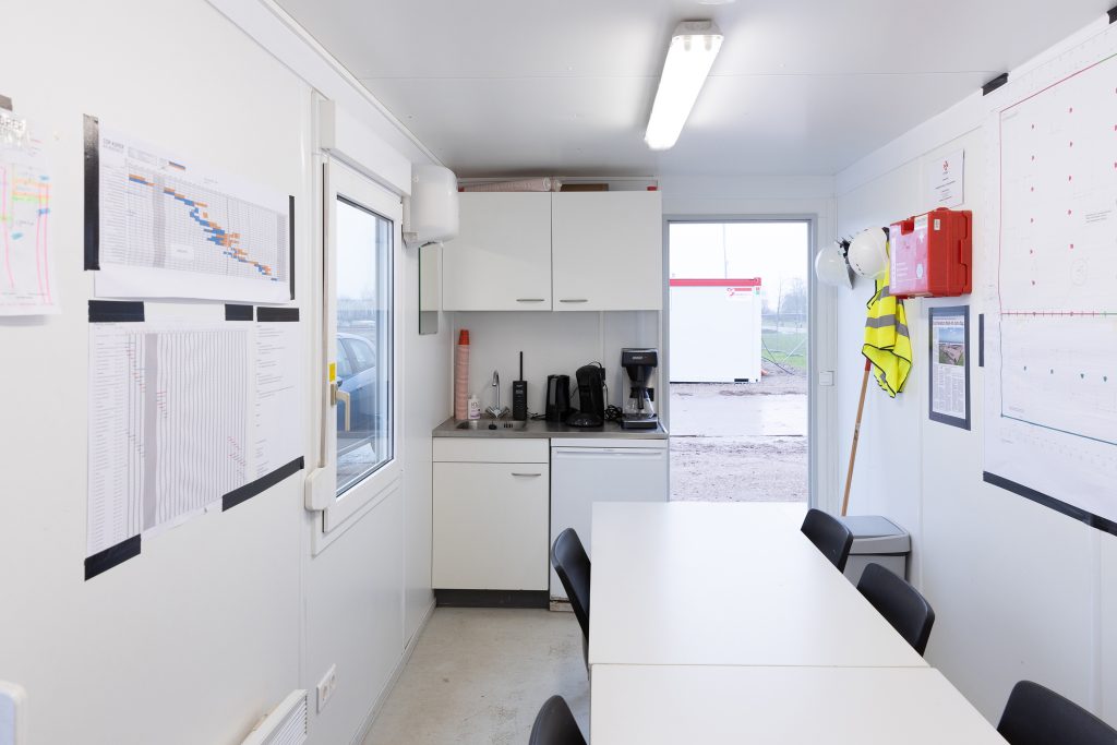 Interieur van een bouwkeet met een keuken, een tafel, tl-verlichting en ehbo-verbanddoos.