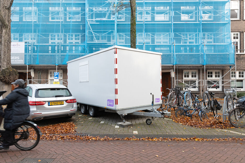 Witte schaftwagen met 4 wielen die geparkeerd staat in een woonwijk, met op de achtergrond een woningblok met steiger ervoor.